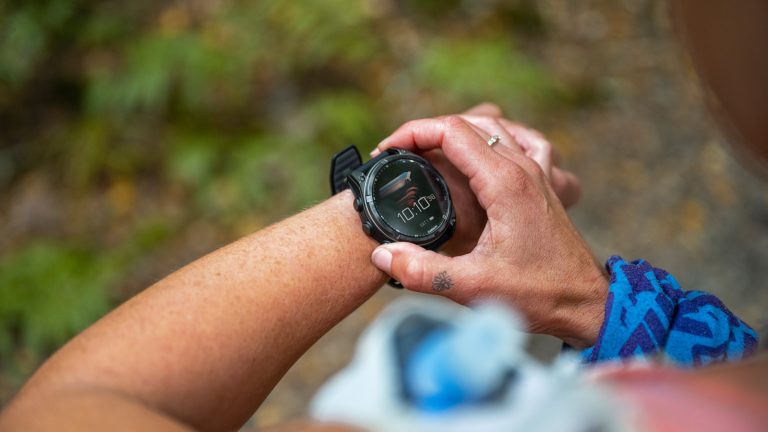 Blick über die Schulter einer Person: Am linken Handgelenk trägt die Person eine Smartwatch. Mit der anderen Hand drückt sie die Knöpfe der Smartwatch.