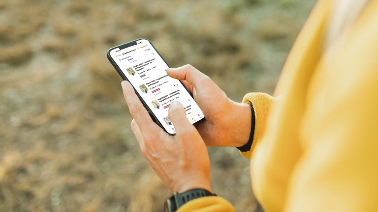 Ein Smartphone mit ge&ouml;ffneter Komoot-App liegt in der Hand einer Person, die eine Smartwatch und eine gelbe Fleecejacke tr&auml;gt.