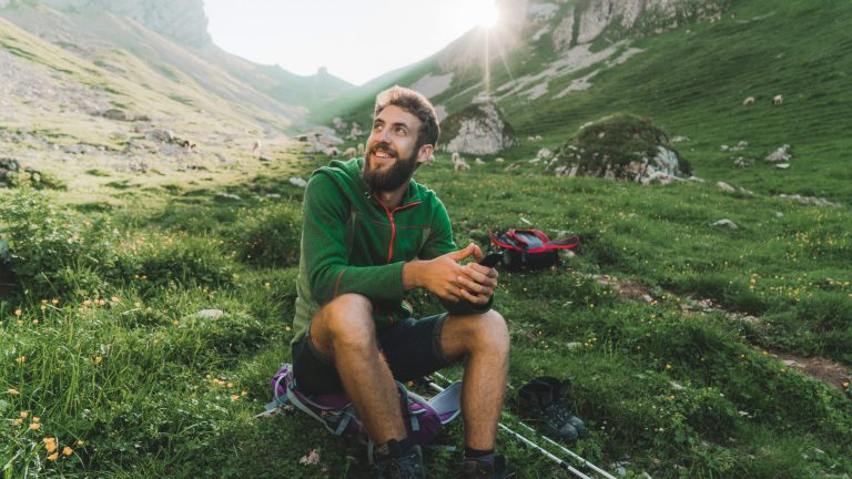 Eine Person macht, auf einem Rucksack sitzend, Rast auf einer Wiese in einer Berglandschaft. Sie h&auml;lt dabei ihr Smartphone in den H&auml;nden.