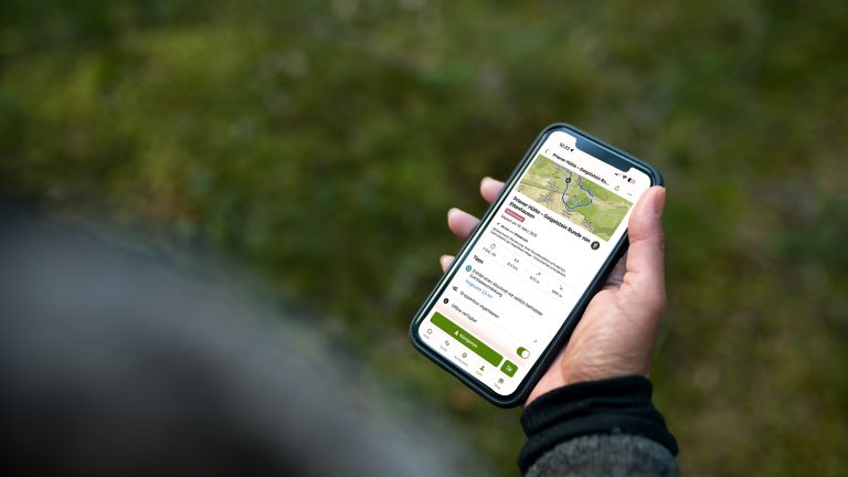 Blick &uuml;ber die Schulter einer Person, die ein Smartphone mit der ge&ouml;ffneten Komoot-App in der Hand h&auml;lt.