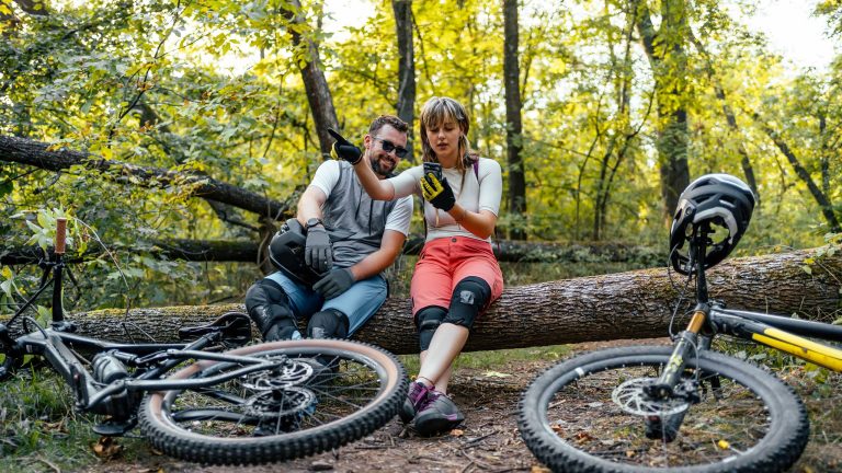 Zwei Personen sitzen auf einem Baumstamm und blicken in ein Smartphone. Ihre Mountainbikes liegen vor ihnen auf dem Weg.&nbsp;