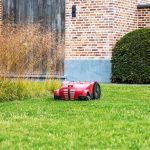 Ein M&auml;hroboter f&auml;hrt &uuml;ber eine Wiese. Im Hintergrund ist eine Hecke und ein Teil eines Ziegelsteinhauses zu sehen.