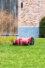 Ein Mähroboter fährt über eine Wiese. Im Hintergrund ist eine Hecke und ein Teil eines Ziegelsteinhauses zu sehen.