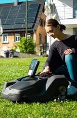 Eine Person kniet hinter einem Mähroboter mit einem geöffneten Gehäuse.