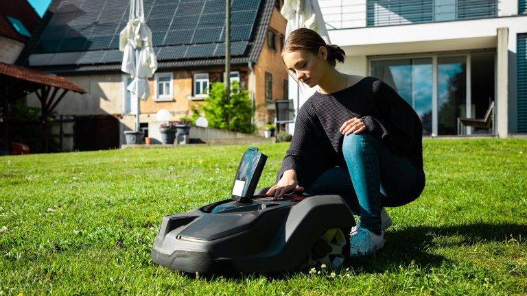 Eine Person kniet hinter einem Mähroboter mit einem geöffneten Gehäuse.