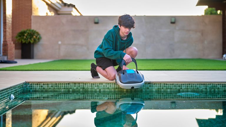 Eine Person kniet am Beckenrand eines Pools in einem Garten und hebt einen Akku-Poolroboter ins Wasser.