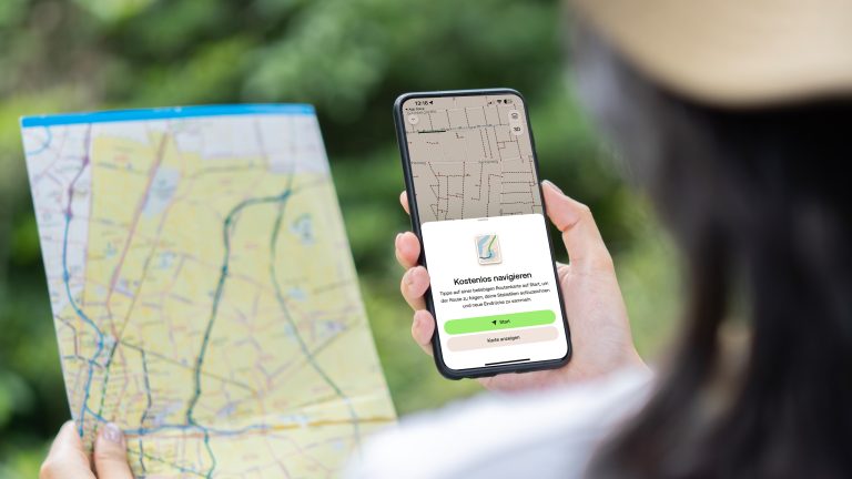 Blick über die Schulter einer Person, die in der linken Hand eine Landkarte hält und in der rechten ein Smartphone mit der geöffneten AllTrails-App.