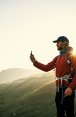Eine Person steht in Wanderkleidung an einer felsigen K&uuml;ste und h&auml;lt ein Smartphone vor sich in der Hand.