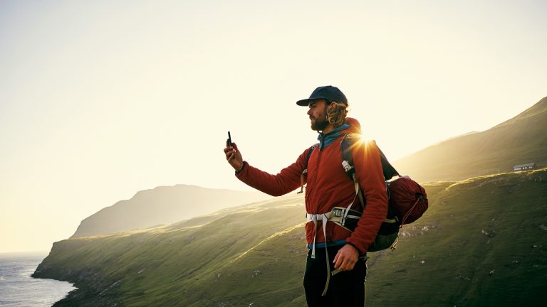 Eine Person steht in Wanderkleidung an einer felsigen Küste und hält ein Smartphone vor sich in der Hand.