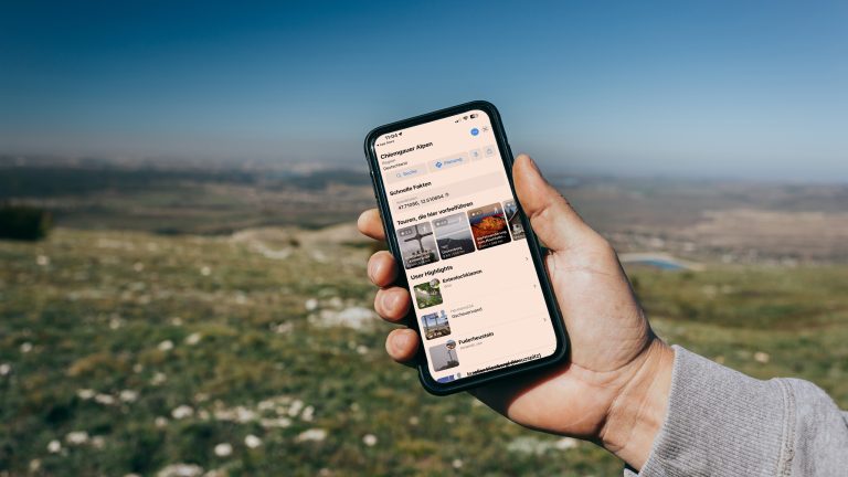 Im Vordergrund steht die Hand einer Person, die ein Smartphone mit der geöffneten Bergfex-App in der Hand hält. Im Hintergrund ist eine hügelige Naturlandschaft zu sehen.