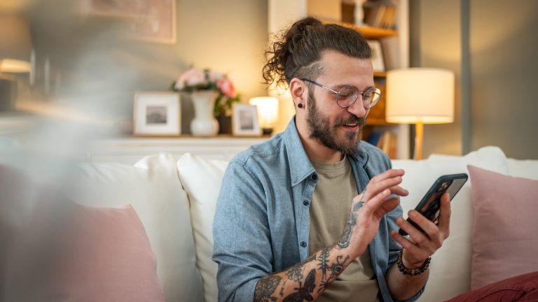 Eine Person mit Bart, langen Haaren und Tattoos sitzt im Wohnzimmer auf der Couch und scrollt auf ihrem Smartphone vor sich in der Hand.