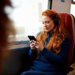 Eine Person sitzt in einem Zug am Fenster, tr&auml;gt Overear-Kopfh&ouml;rer und schaut auf ihr Smartphone.