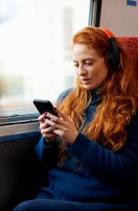 5G Handys, Was können sie? Eine Person sitzt in einem Zug am Fenster, trägt Overear-Kopfhörer und schaut auf ihr Smartphone.