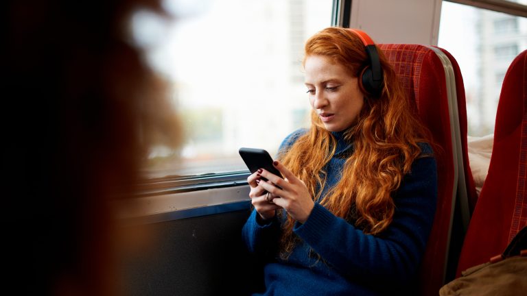 Eine Person sitzt in einem Zug am Fenster, trägt Overear-Kopfhörer und schaut auf ihr Smartphone.