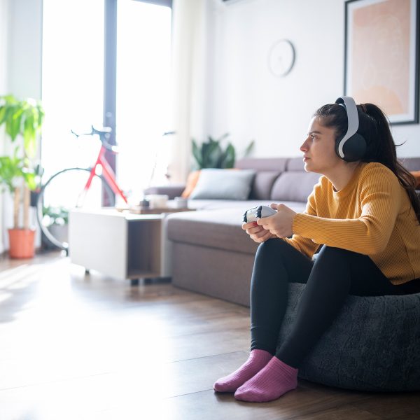 Eine Person auf dem Sofa und schaut konzentriert auf einen Fernseher. Sie trägt Over-Ear-Kopfhörer und hält einen Controller in den Händen.