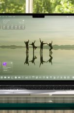 Ein aufgeklapptes MacBook auf einem Glasschreibtisch vor einem Fenster. Auf dem Screen der Startbildschirm unter macOS 26.