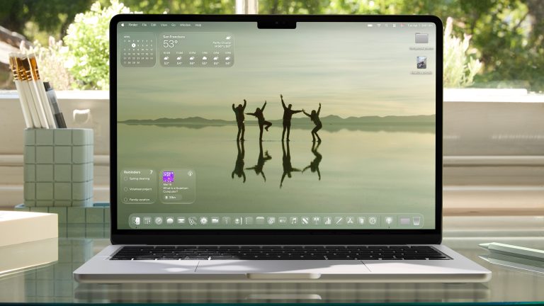 Ein aufgeklapptes MacBook auf einem Glasschreibtisch vor einem Fenster. Auf dem Screen der Startbildschirm unter macOS 26.