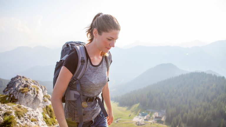 Seitansicht einer Person in Jeansshorts und mit Wanderrucksack. Im Hintergrund ist alpine Landschaft zu sehen.