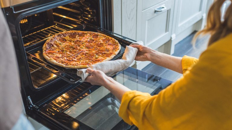 Moderne Backöfen, Systeme & Modelle Eine Person holt mit einem zusammengerollten Küchenhandtuch ein Blech mit einer Pizza aus einem Ofen in einer Küche.