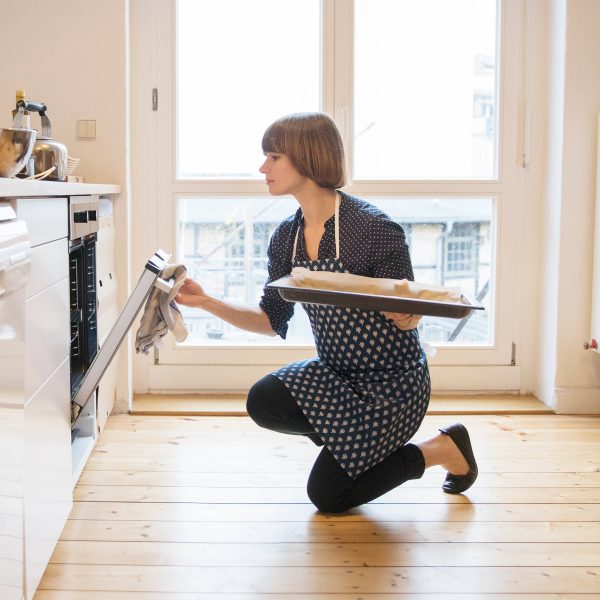 Eine Person &ouml;ffnet mit einem Backblech in der Hand kniend einen Ofen in einer K&uuml;che.