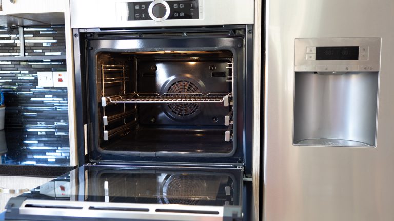 Moderne Backöfen, Systeme & Modelle Ein geöffneter Backofen, der in einer Küche neben einem Kühlschrank auf Oberkörperhöhe verbaut ist.