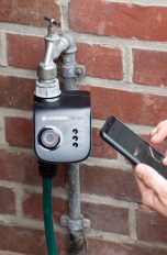 Eine Person kniet neben einem Wasseranschluss, an dem ein Gardena Smart-System angeschlossen ist und bedient ihr Smartphone.