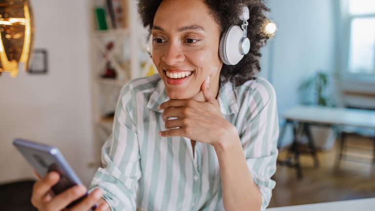 Eine Person mit Overhead-Kopfhörern sitzt in an einem Tisch und schaut auf ihr Smartphone.