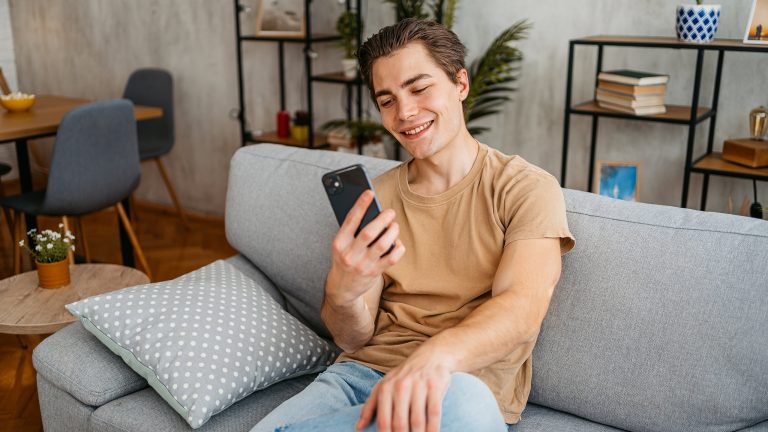 Eine Person sitzt auf einem Sofa in einem Wohnzimmer und blickt auf ihr Smartphone. 