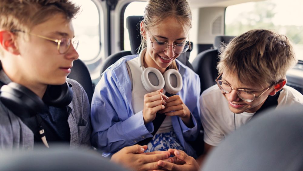 Drei Jugendliche sitzen nebeneinander auf der Rückbank eines Autos. Die beiden äußeren spielen in der Mitte mit ihren Händen ein Spiel. Alle tragen Kopfhörer um ihren Hals.