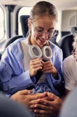 Drei Jugendliche sitzen nebeneinander auf der Rückbank eines Autos. Die beiden äußeren spielen in der Mitte mit ihren Händen ein Spiel. Alle tragen Kopfhörer um ihren Hals.