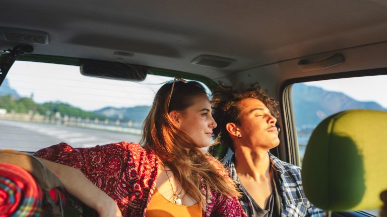 Zwei Personen auf der R&uuml;ckbank eines Autos gucken aus dem Fenster in die Sonne. Ihre Haare fliegen im Wind.