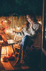 Praktische Balkon-Gadgets für Frühling und Sommer Eine Person sitzt im Dunkeln auf ihrem gemütlich gestalteten Balkon, hält ein Weinglas in der Hand und sieht lächelnd in ihren Laptop. Im Hintergrund ist eine Lichterkette zu sehen.