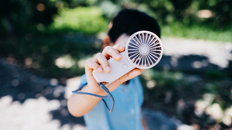 Im Fokus steht die R&uuml;ckansicht eines mobilen Mini-Ventilators, den eine Person in blauem Shirt in den H&auml;nden h&auml;lt.