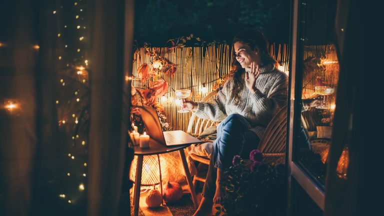 Eine Person sitzt im Dunkeln auf ihrem gem&uuml;tlich gestalteten Balkon, h&auml;lt ein Weinglas in der Hand und sieht l&auml;chelnd in ihren Laptop. Im Hintergrund ist eine Lichterkette zu sehen.