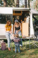 Elektrische Kühlboxen, Modelle, Technik & Tipps für unterwegs Zwei Personen und ein Kind stehen vor einem Camper auf einem Campingplatz. Auf dem Campingtisch bereiten sie gerade Essen vor.