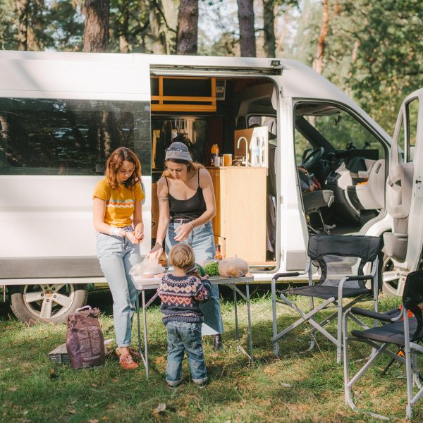 Zwei Personen und ein Kind stehen vor einem Camper auf einem Campingplatz. Auf dem Campingtisch bereiten sie gerade Essen vor.