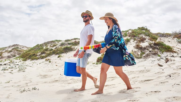 Zwei Personen laufen in Strandkleidung &uuml;ber sandige D&uuml;nen. In den H&auml;nden tragen sie einen Sonnenschirm und eine K&uuml;hlbox.