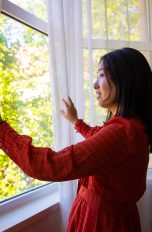 LuftqualiLuftqualität verbessern, Durchatmen für Allergiker*innen taet-5 Eine Person steht an einem geöffneten Fenster. Sie öffnet den Vorhang, der vor dem Fenster hängt, mit beiden Händen.