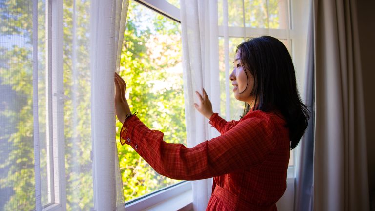 Eine Person steht an einem ge&ouml;ffneten Fenster. Sie &ouml;ffnet den Vorhang, der vor dem Fenster h&auml;ngt, mit beiden H&auml;nden.