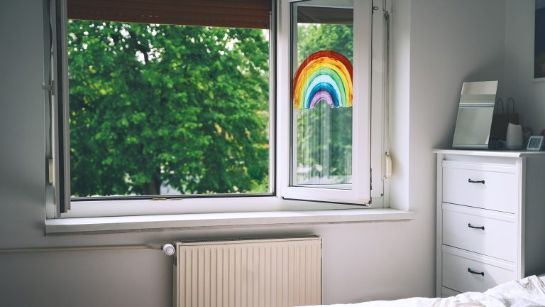 In einem Kinderzimmer ist das Fenster weit ge&ouml;ffnet. An einer Seite des Fensters klebt ein Regenbogen. Vor dem Fenster sind B&auml;ume zu sehen.