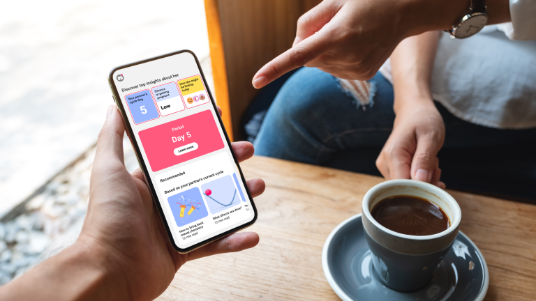 Male’s Flo for Partners Eine Person sitzt an einem Tisch mit einer Kaffeetasse. In den Händen hält sie ein Smartphone, auf dem die Flo-App geöffnet ist.