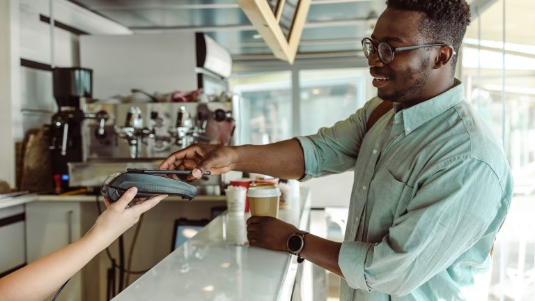 Eine Person steht am Tresen eines Cafés. In der linken Hand hält sie einen Kaffeebecher, in der rechten das Smartphone, mit dem sie gerade bezahlt.
