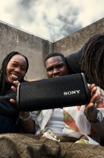 Vier Personen sitzen zusammen und lachen. Eine Person h&auml;lt einen Bluetooth-Lautsprecher in den H&auml;nden.