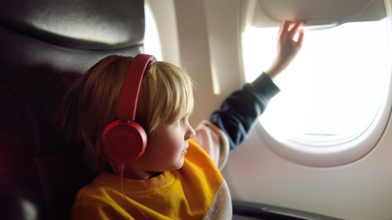 Ein Kind sitzt in einem Flugzeugsitz, tr&auml;gt Over-Ear-Kopfh&ouml;rer und schlie&szlig;t gerade die Blende des Flugzeugfensters.
