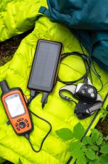 Mit diesen 10 Urlaubs-Gadgets kann der Sommer kommen Ein Satellitenkommunikationsgerät liegt auf einem neongrünen Schlafsack. Daneben liegt ein dunkelblauer Outdoor-Rucksack.