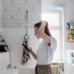 Eine Person steht im Badezimmer vor dem Spiegel. Mit der linken Hand fasst sie sich in die Haare, in der rechten Hand hält sie ein Smartphone.