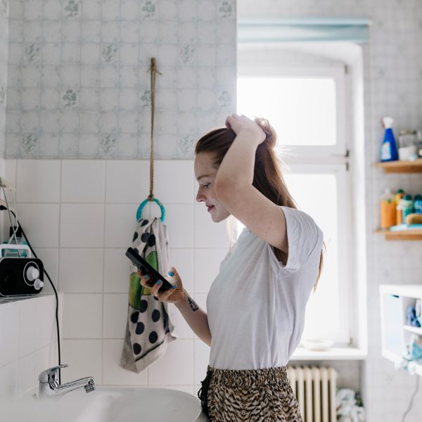Eine Person steht im Badezimmer vor dem Spiegel. Mit der linken Hand fasst sie sich in die Haare, in der rechten Hand h&auml;lt sie ein Smartphone.