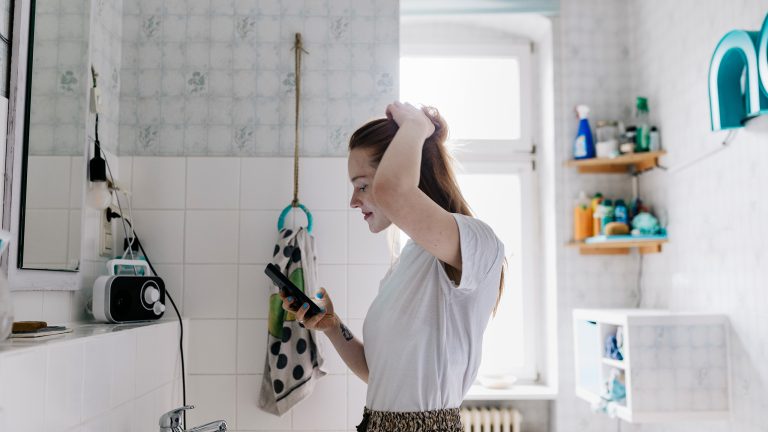 Eine Person steht im Badezimmer vor dem Spiegel. Mit der linken Hand fasst sie sich in die Haare, in der rechten Hand h&auml;lt sie ein Smartphone.