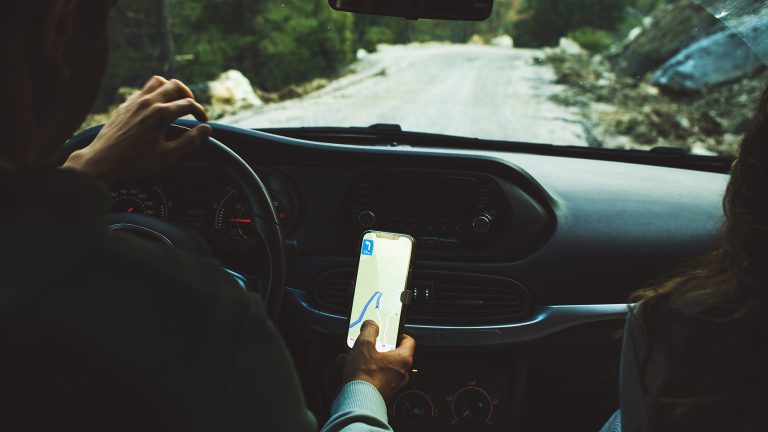 Eine Person von hinten auf dem Fahrersitz eines Autos. Sie f&auml;hrt auf einer Stra&szlig;e, w&auml;hrend ein Handy an der Konsole die Route anzeigt.