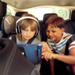 Zwei Kinder auf der Rückbank eines Autos schauen zusammen auf ein Tablet. Ein Kind trägt Kopfhörer.